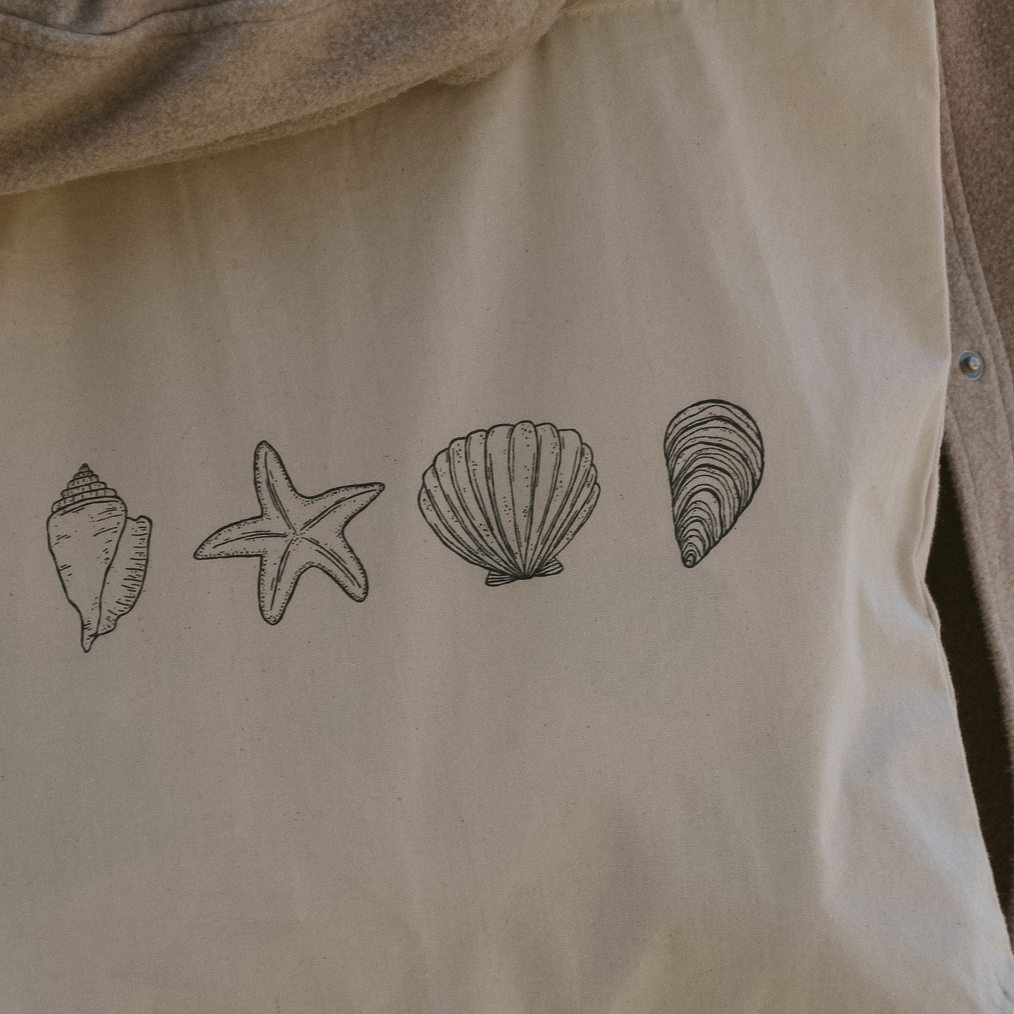 Nahaufnahme einer naturfarbenen Stofftasche mit vier schwarz illustrierten maritimen Motiven: Muschel, Seestern, Jakobsmuschel und Schneckenhaus.