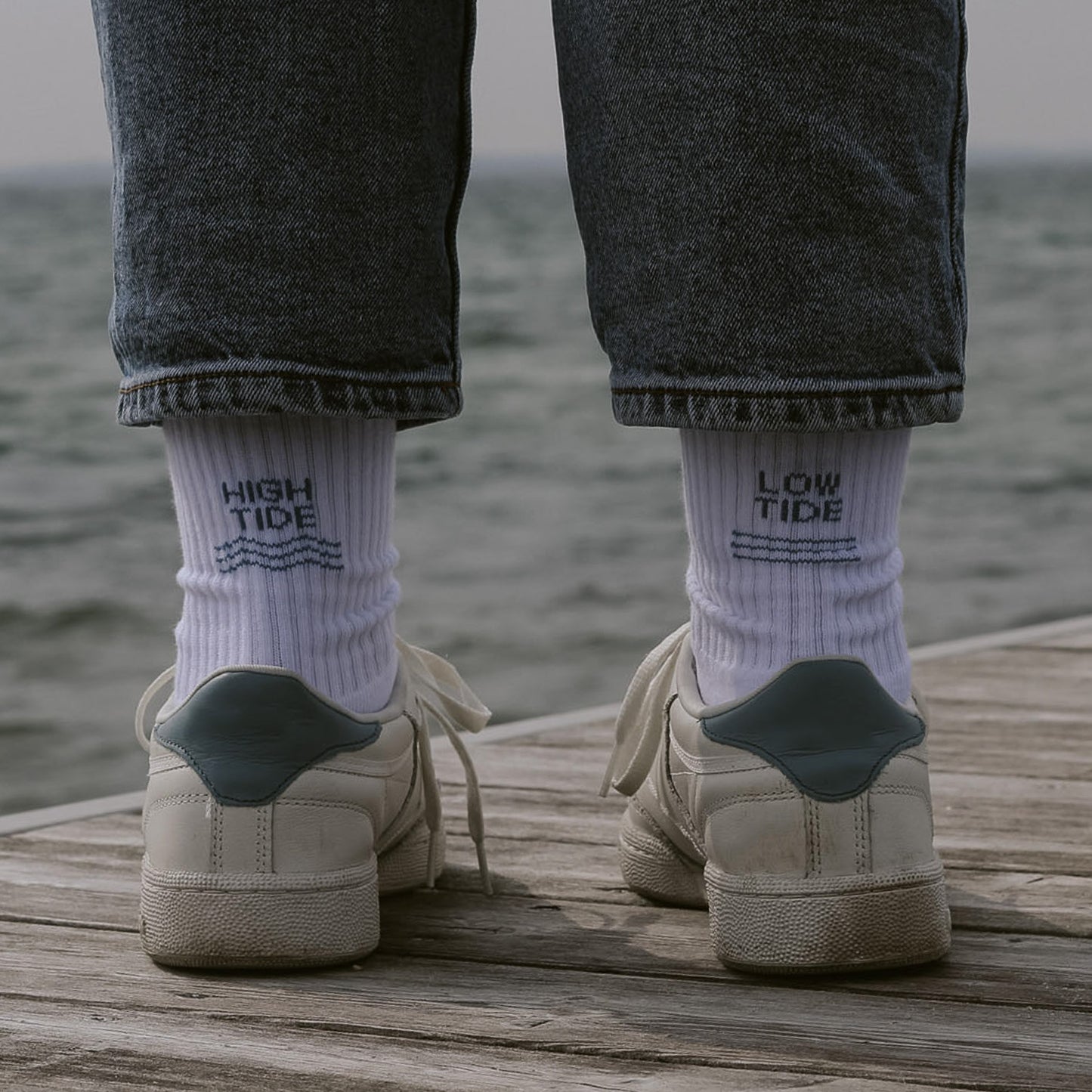Person von hinten mit weißen Saltwaterdesigns Tennissocken, High Tide links, Low Tide rechts – getragen mit weißen Sneakern auf einem Holzsteg mit Meer im Hintergrund.