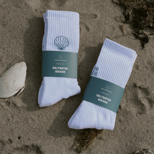 Saltwater Designs Socken Bundle – Muschel und High Tide Low Tide, beide fertig verpackt mit petrolblauer Banderole, nebeneinander auf Sand liegend mit einer Muschel daneben.