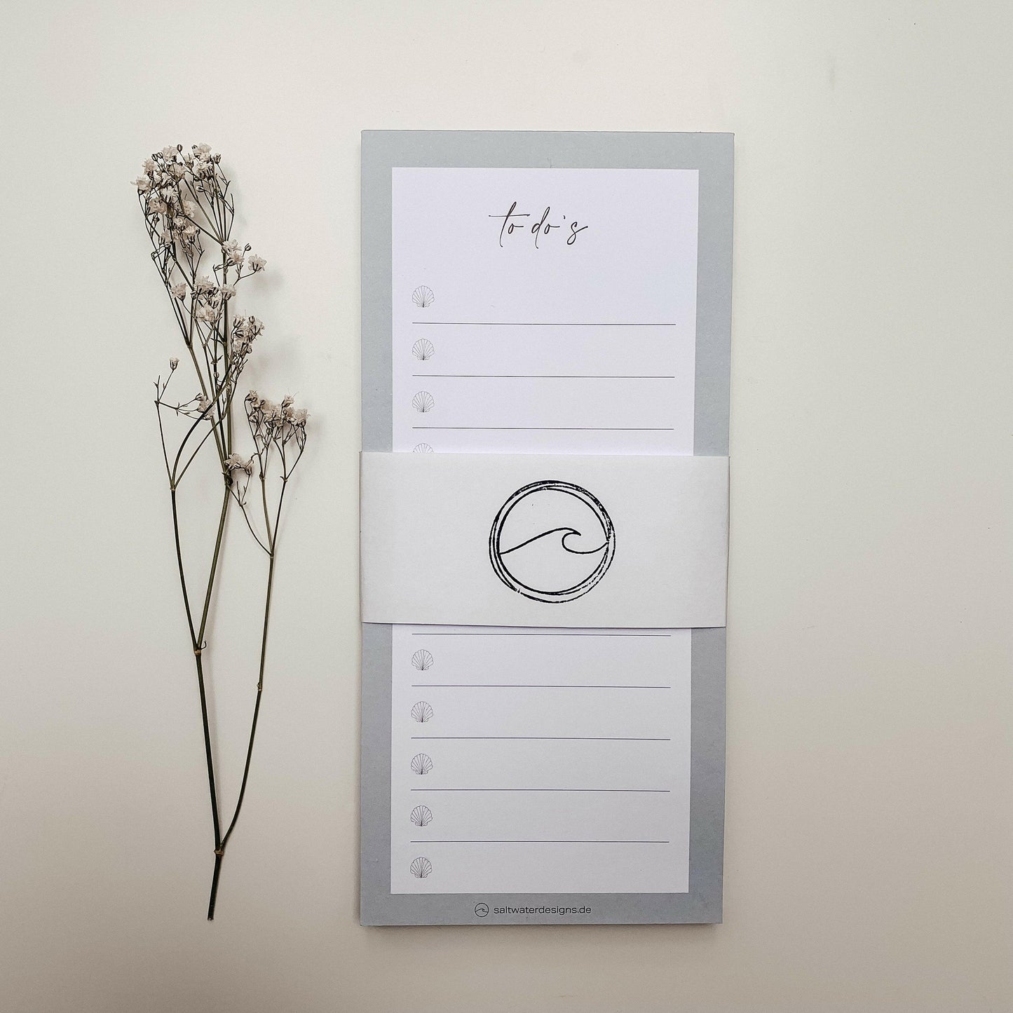 Flach aufgenommener Notizblock im Hochformat mit hellgrauem Rand und der Aufschrift 'to do's' oben. Die Seiten sind mit kleinen Muschelsymbolen und Linien zum Beschriften versehen. Um den Block liegt ein weißes Papierband mit schwarzem Wellenlogo. Links daneben liegen zwei getrocknete Zweige mit kleinen Blüten auf hellem Hintergrund. Unten am Blockrand ist die Webadresse 'saltwaterdesigns.de' zu sehen.