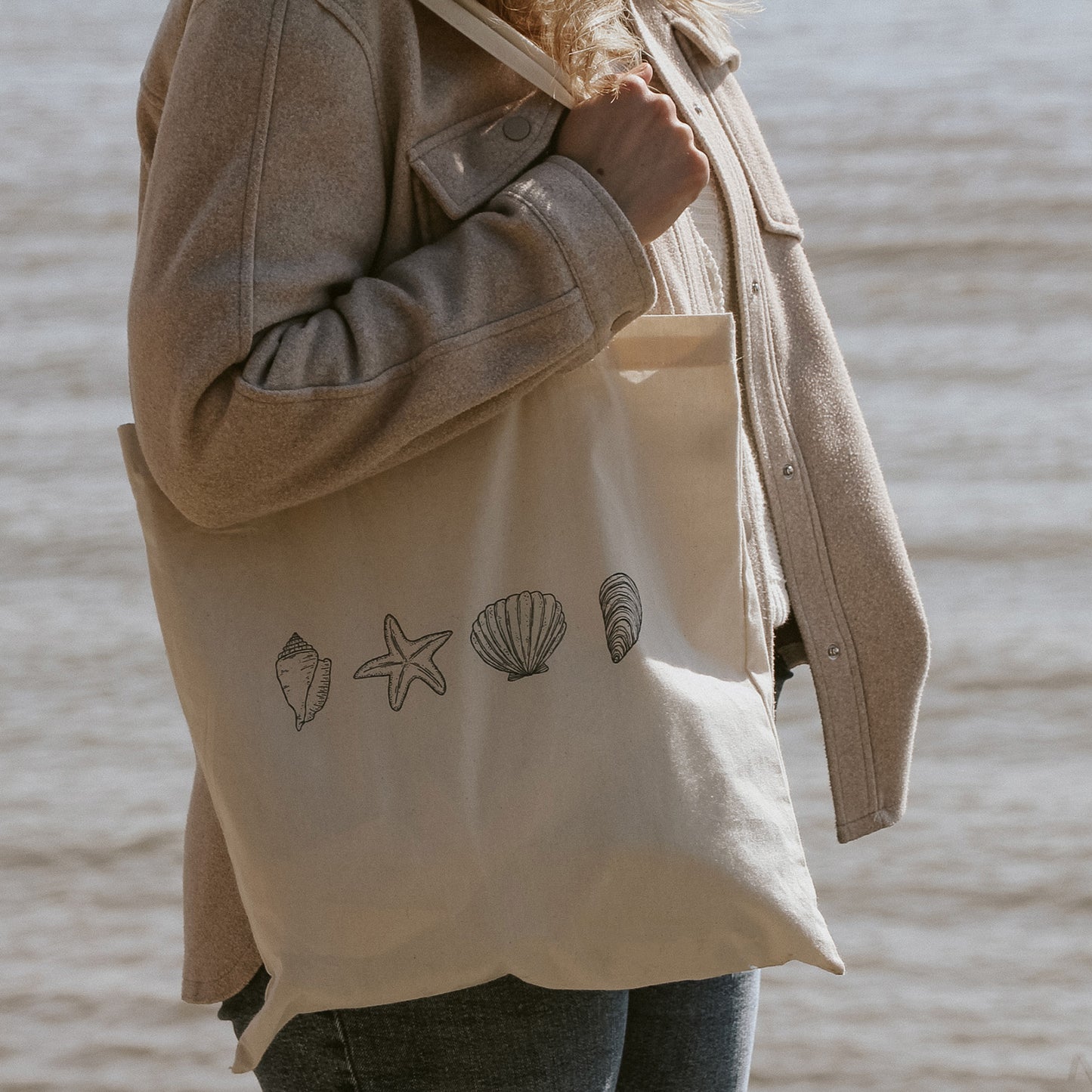Frau am Meer mit naturfarbener Stofftasche aus Baumwolle, bedruckt mit maritimen Illustrationen von Muscheln und Seestern.