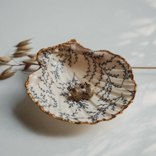 Handbemalte Jakobsmuschel mit goldfarbenem Rand und dunkelblauem Blattmuster im Inneren, darin liegend ein Paar goldfarbener Muschelohrringe, dekoriert mit getrockneten Grashalmen im Hintergrund.