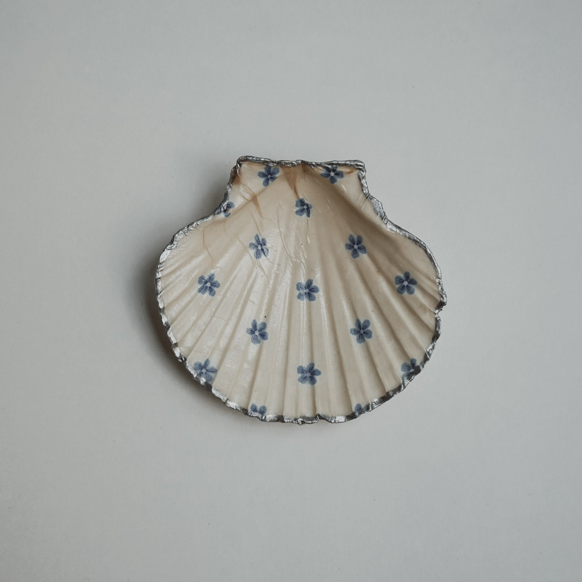 Handbemalte Jakobsmuschel mit cremefarbenem Hintergrund, verziert mit kleinen blauen Blumenmustern und silbernem Rand, auf hellem Untergrund platziert.