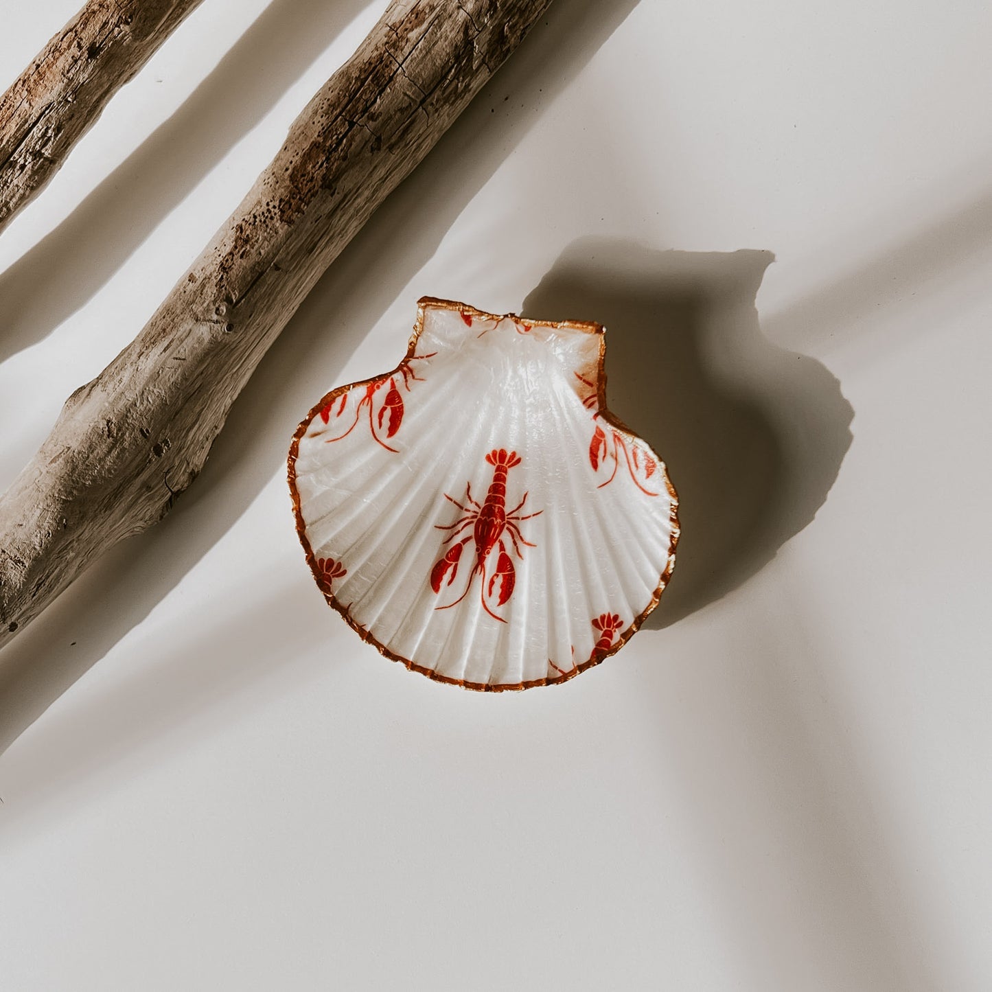 Dekorative weiße Muschelschale mit rotem Hummer-Design und vergoldetem Rand, fotografiert in natürlichem Licht neben Treibholz.