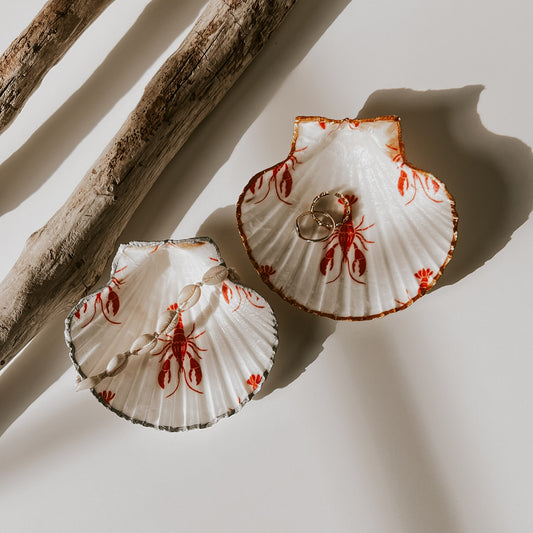 Zwei dekorative Muschelschalen mit rotem Hummer-Design eine mit goldfarbenem Rand, eine mit silberfarbenem Rand, gefüllt mit Schmuck, arrangiert neben Treibholz.