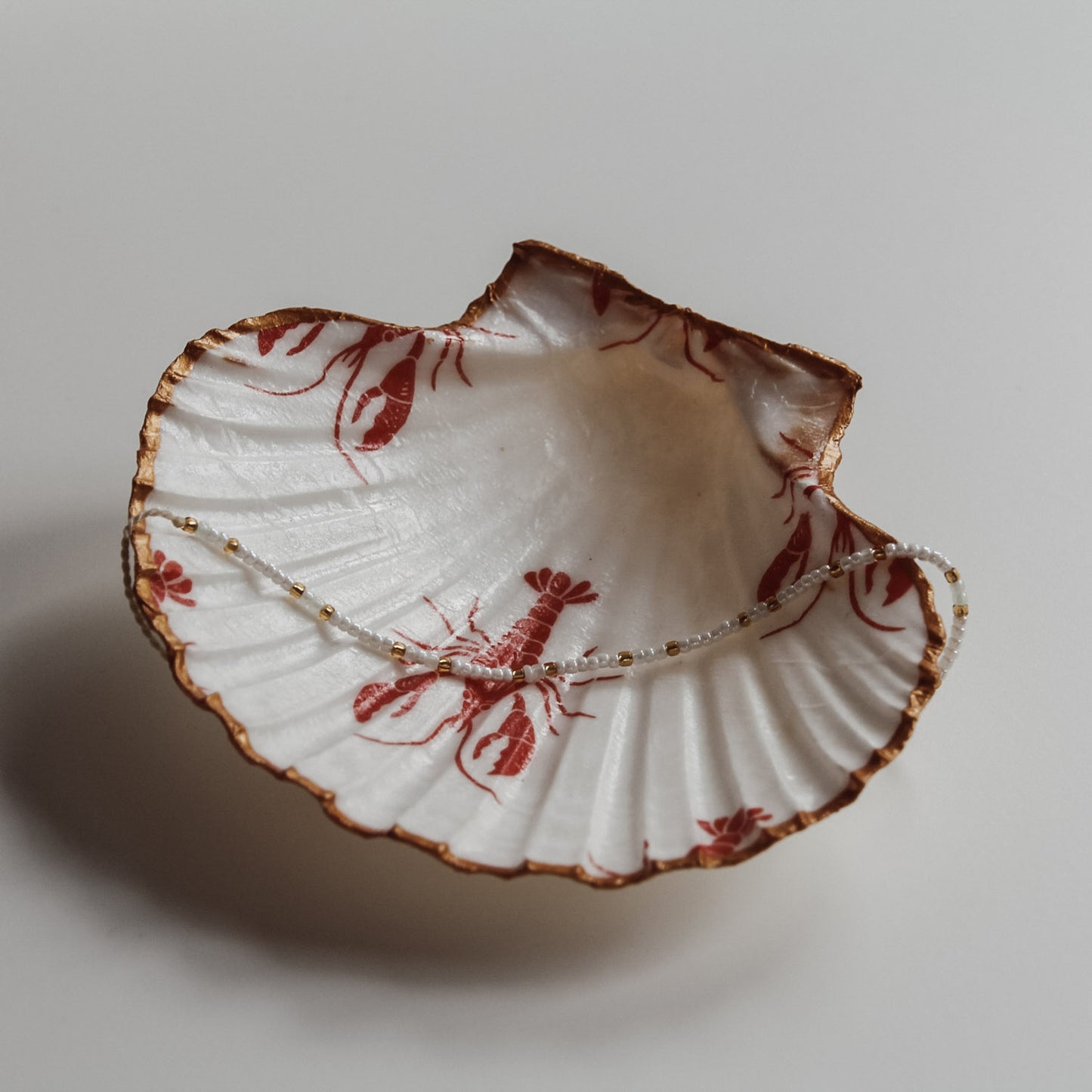 Dekorative weiße Muschelschale mit rotem Hummer-Design und vergoldetem Rand, fotografiert in natürlichem Licht neben Treibholz. In der Schale liegt ein Perlenarmband und das soll zeigen, dass sie sich auch als Schmuckschale nutzen lässt.