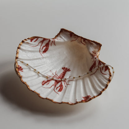 Dekorative weiße Muschelschale mit rotem Hummer-Design und vergoldetem Rand, fotografiert in natürlichem Licht neben Treibholz. In der Schale liegt ein Perlenarmband und das soll zeigen, dass sie sich auch als Schmuckschale nutzen lässt.