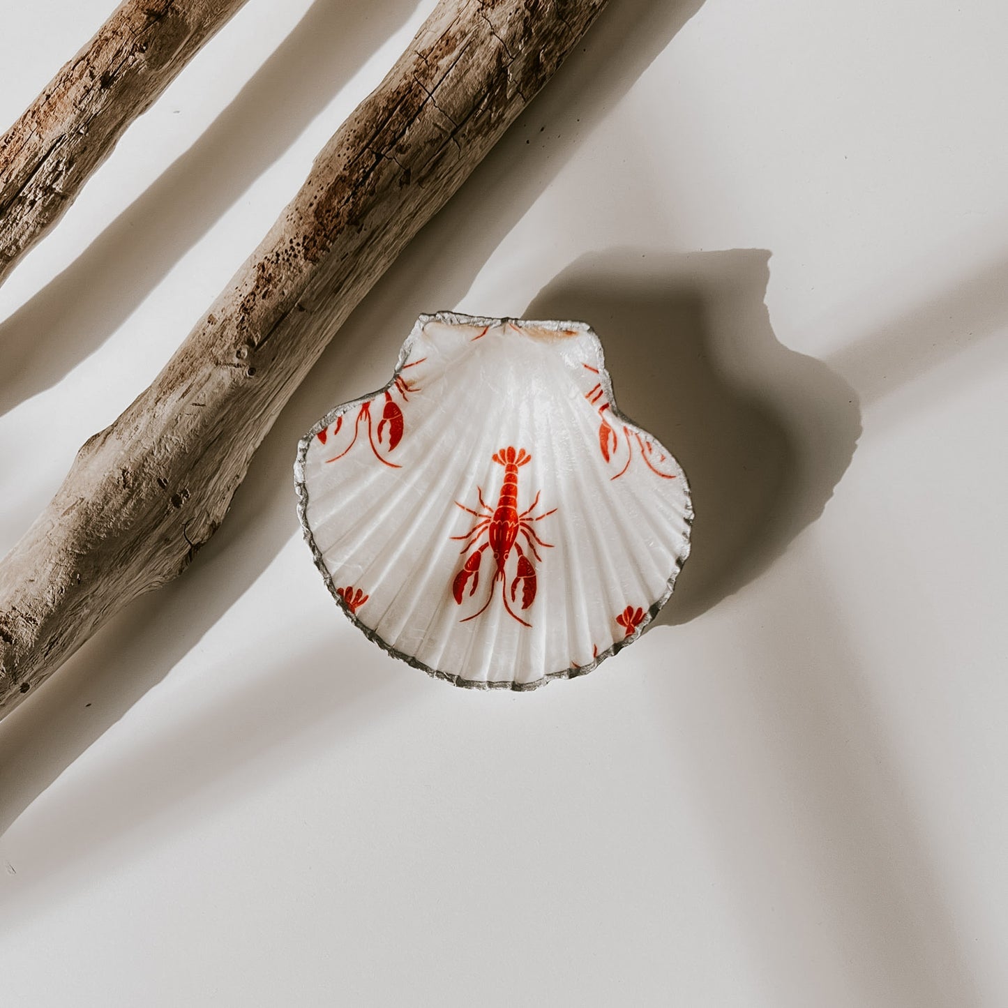 Dekorative weiße Muschelschale mit rotem Hummer-Design und silberfarbenem Rand, fotografiert in natürlichem Licht neben Treibholz.