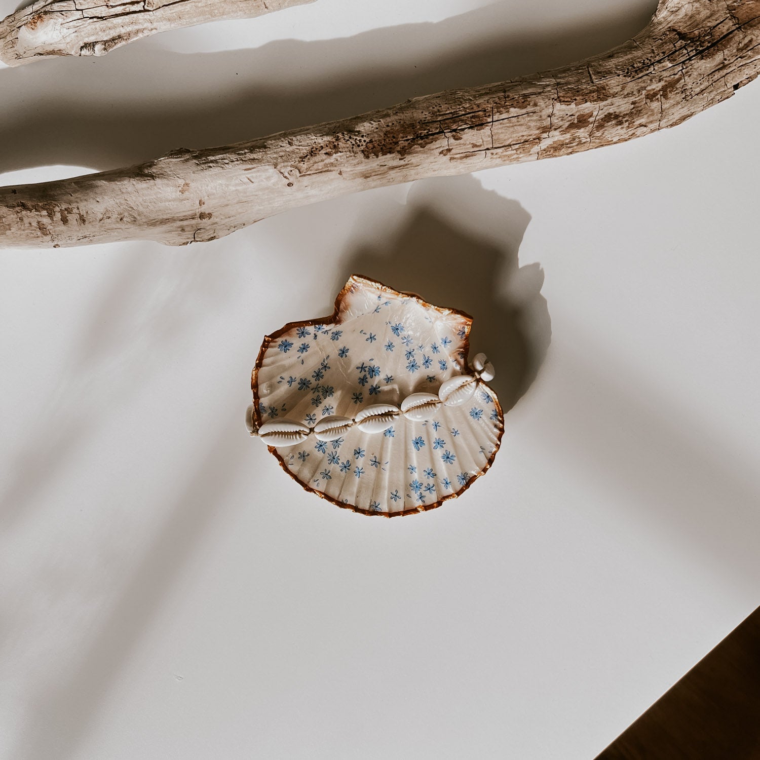 Dekorative weiße Muschelschale mit Blumen-Design und vergoldetem Rand, fotografiert in natürlichem Licht neben Treibholz. In der Schale liegt ein Muschelarmband.