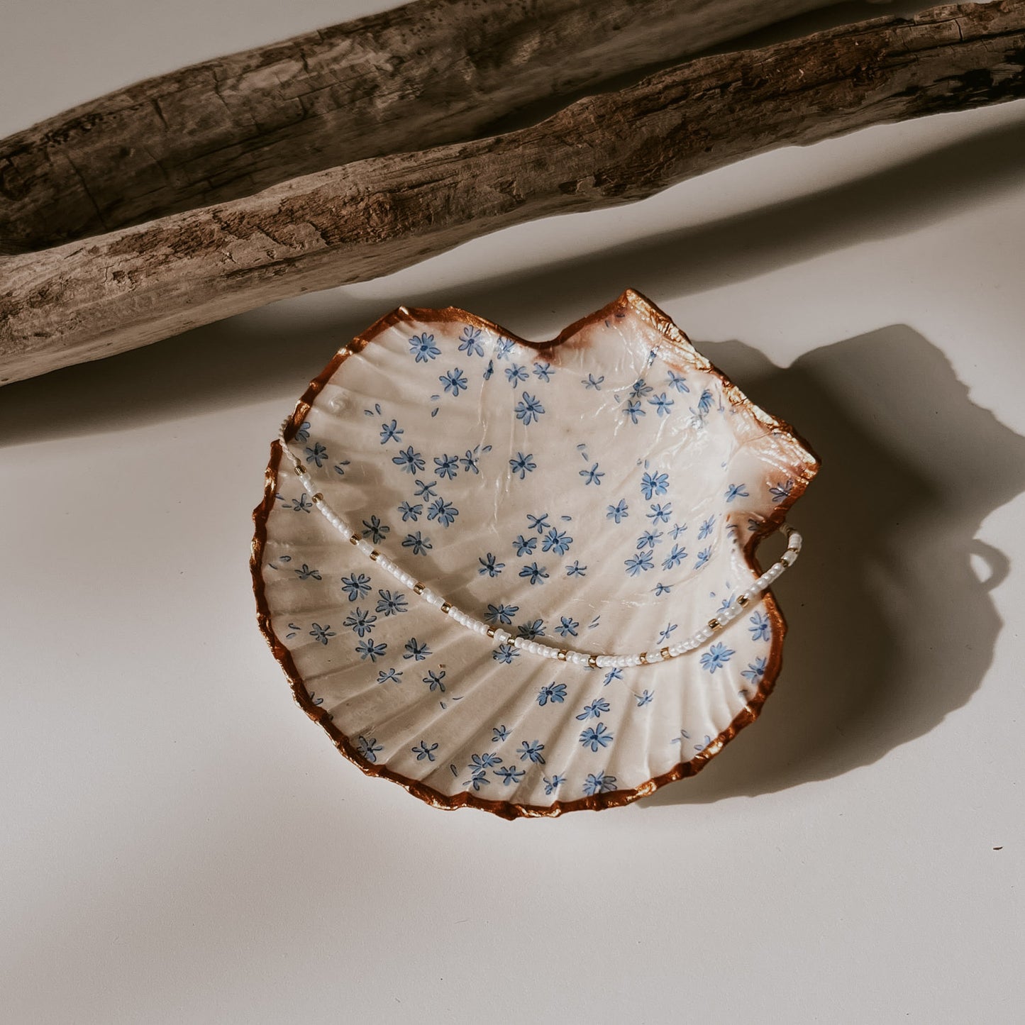 Dekorative weiße Muschelschale mit Blumen-Design und vergoldetem Rand, in der Schale liegt ein Perlenarmband, sie ist fotografiert in natürlichem Licht neben Treibholz