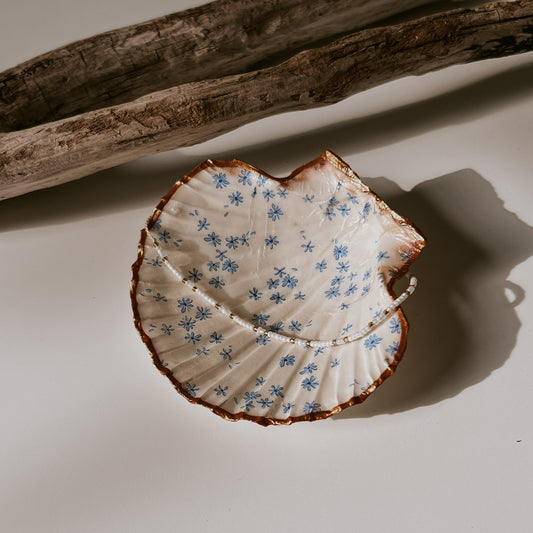 Dekorative weiße Muschelschale mit Blumen-Design und vergoldetem Rand, in der Schale liegt ein Perlenarmband, sie ist fotografiert in natürlichem Licht neben Treibholz