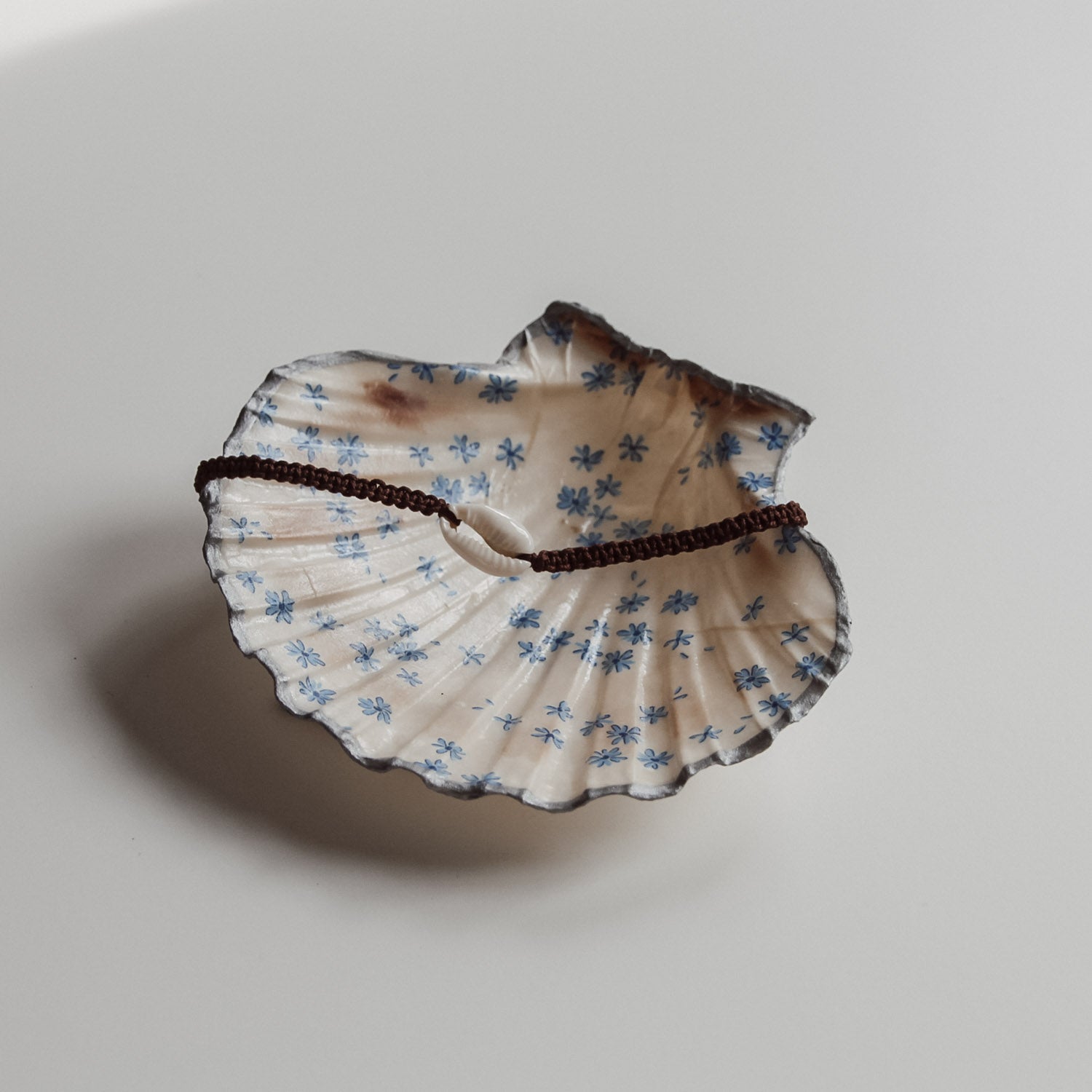 Schlichte Muschelschale mit blauem Blumen Design und silberfarbenem Rand, gefüllt mit braunem Muschelarmband. Handgemacht von saltwaterdesigns.