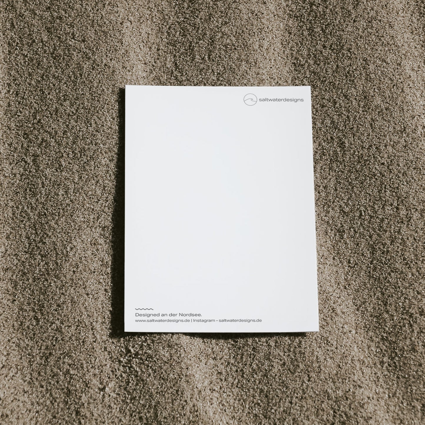 Schlichte Aufnahme von der Postkartenrückseite einer saltwaterdesigns-Postkarte auf dem Sand.