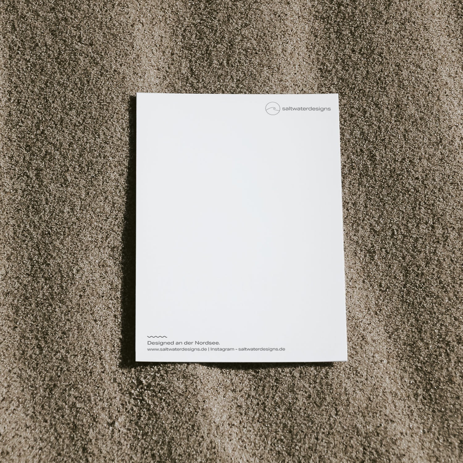Schlichte Aufnahme von der Postkartenrückseite einer saltwaterdesigns-Postkarte auf dem Sand.