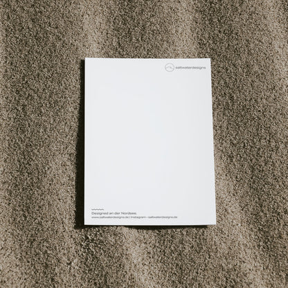 Schlichte Aufnahme von der Postkartenrückseite einer saltwaterdesigns-Postkarte auf dem Sand.