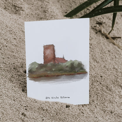 Nahaufnahme einer Postkarte mit der Illustration der „Alten Kirche Pellworm“, Karte ist eingesteckt in Sand.