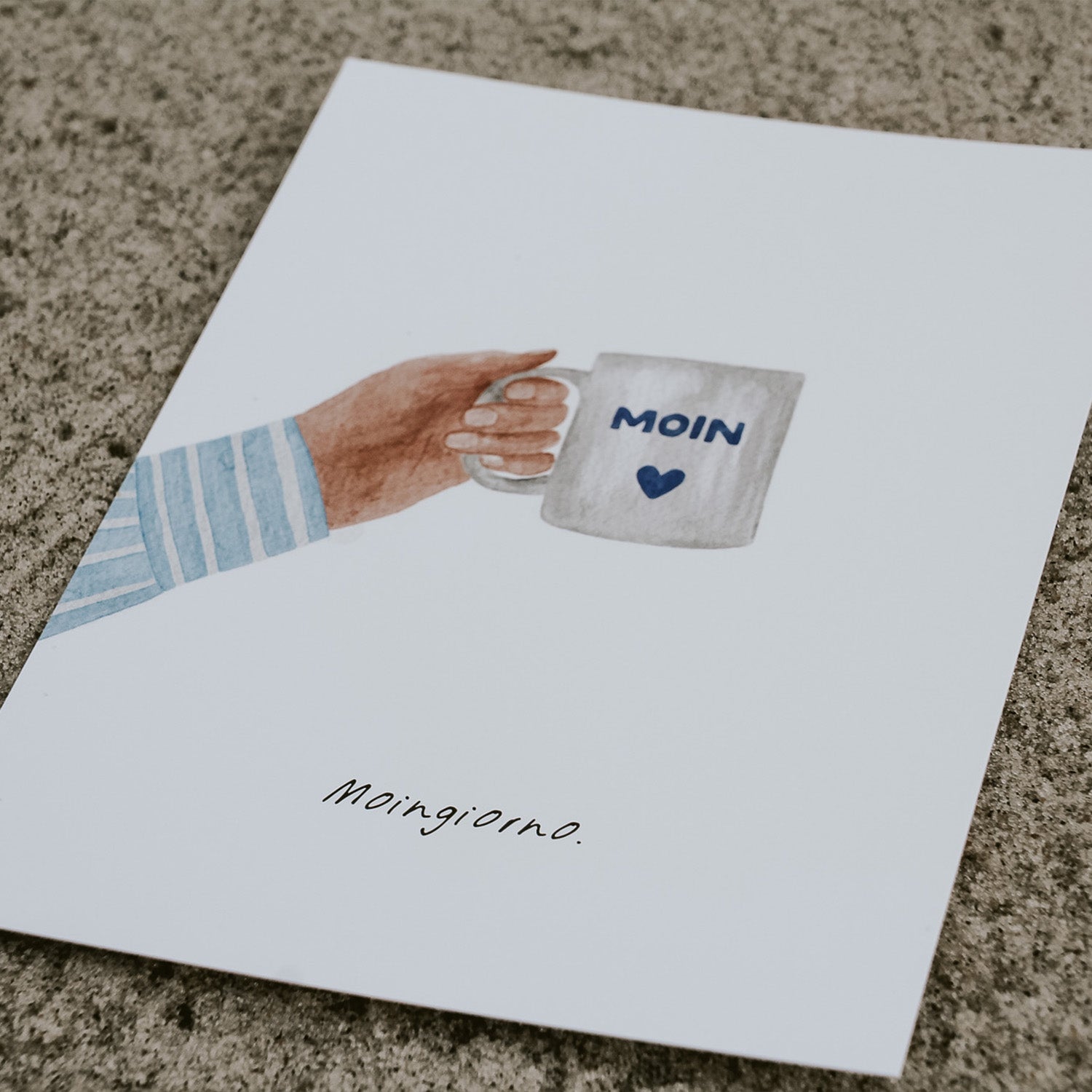 Detailansicht der Postkarte für Kaffeeliebhaber: Aquarellillustration einer Hand mit 'Moin'-Tasse, darunter der Spruch'Moingiorno'. Die Karte liegt auf Sand und versprüht norddeutsche Gelassenheit. 