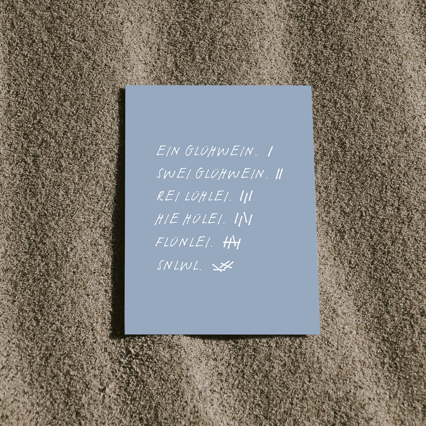 Blaue Postkarte mit lustigem Spruch "Glühwein", liegt auf sandigem Untergrund.
