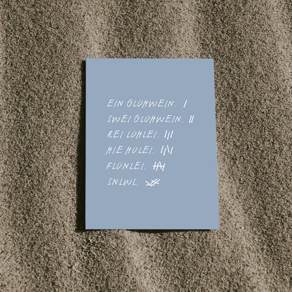 Blaue Postkarte mit lustigem Spruch "Glühwein", liegt auf sandigem Untergrund.