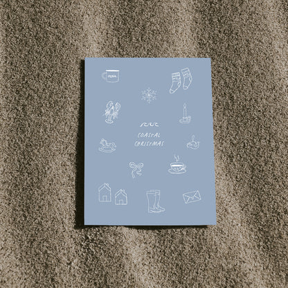 Blaue Postkarte mit dem Schriftzug „Coastal Christmas“ und nordischen, maritimen Illustrationen, die auf sandigem Untergrund liegt.