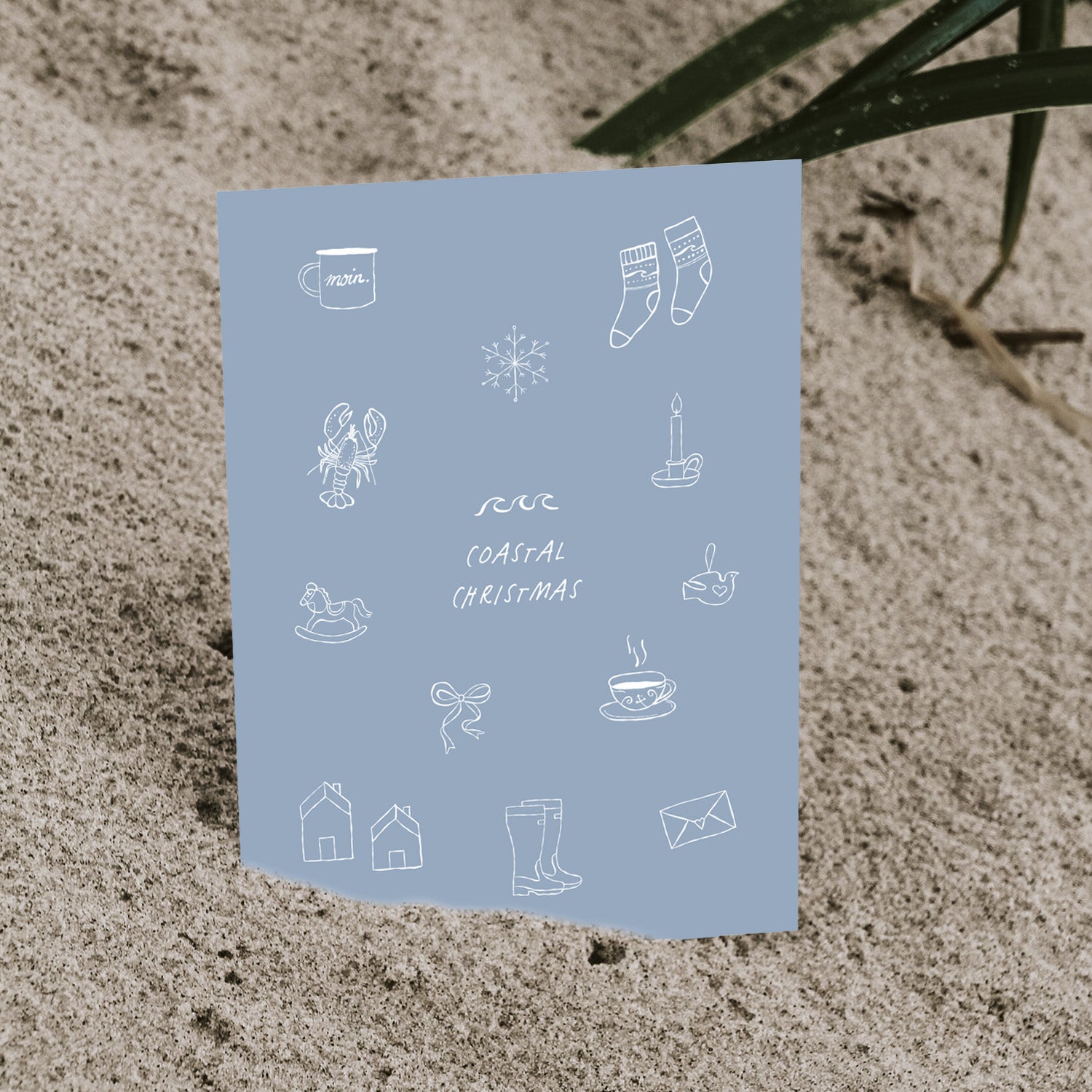 Blaue „Coastal Christmas“ Postkarte von saltwaterdesigns mit nordischen, maritimen Illustrationen, leicht im Sand eingesteckt.