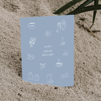 Blaue „Coastal Christmas“ Postkarte von saltwaterdesigns mit nordischen, maritimen Illustrationen, leicht im Sand eingesteckt.