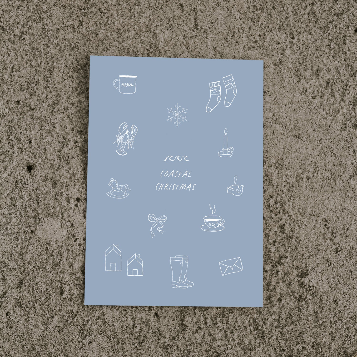 Draufsicht auf die blaue „Coastal Christmas“ Postkarte von saltwaterdesigns mit nordischen, maritimen Illustrationen, liegend auf sandigem Untergrund.