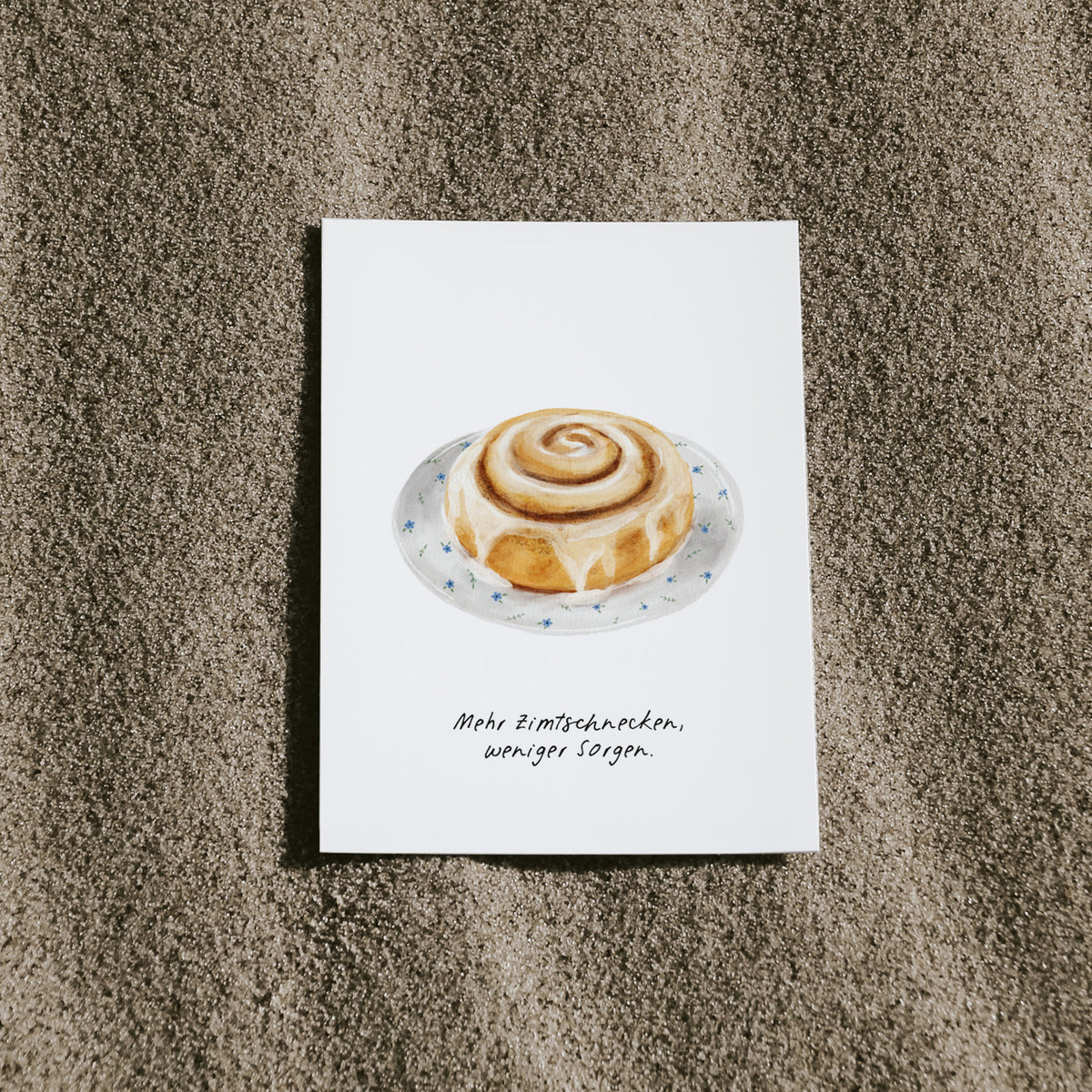 Postkarte Zimtschnecke von saltwaterdesigns liegend auf einem Sandstrand.