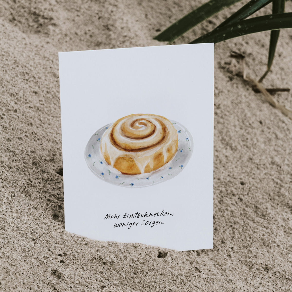 Postkarte Zimtschnecke mit dem Spruch "Mehr Zimtschnecken, weniger Sorgen" im Sand stehend, im Hintergrund Strandgras. By saltwaterdesigns.