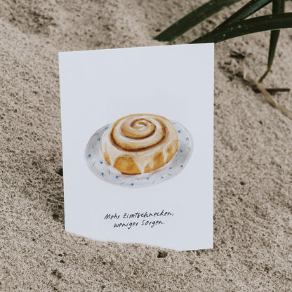 Postkarte Zimtschnecke mit dem Spruch "Mehr Zimtschnecken, weniger Sorgen" im Sand stehend, im Hintergrund Strandgras. By saltwaterdesigns.