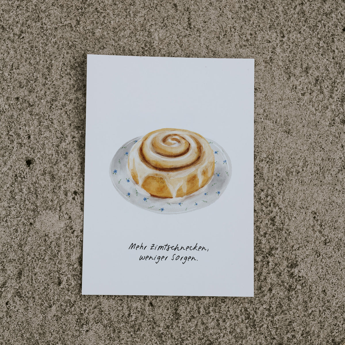 Postkarte Zimtschnecke von saltwaterdesigns liegend auf hellem Sand. Für Zimtschneckenfans