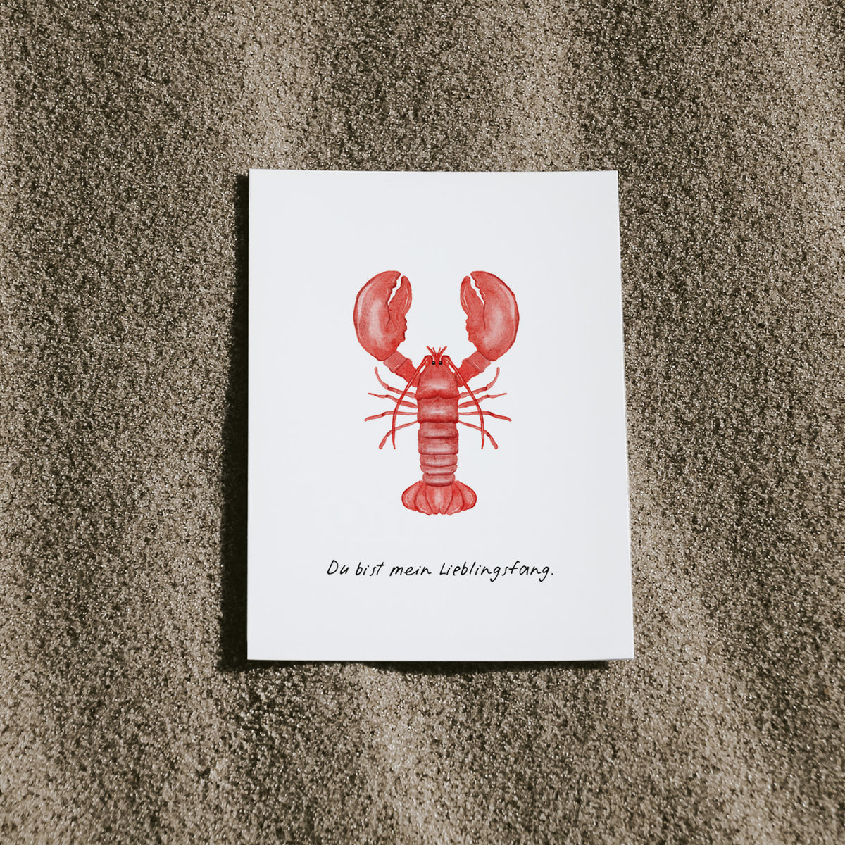 Postkarte Hummer von saltwaterdesigns liegend auf einem Sandstrand.