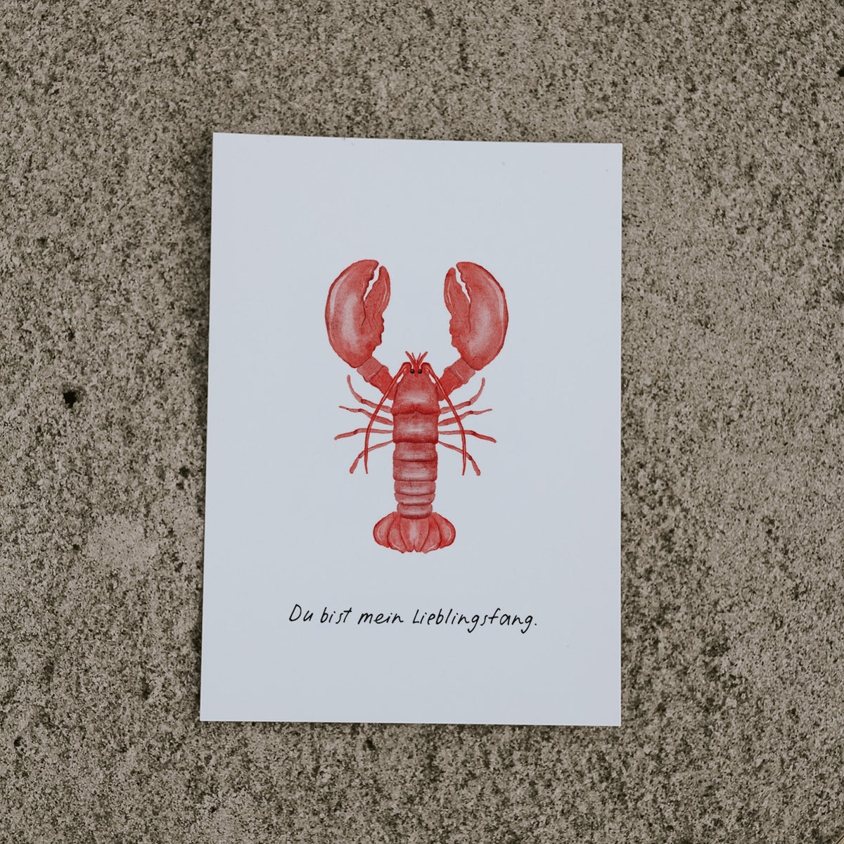 Postkarte Hummer von saltwaterdesigns liegend auf hellem Sanduntergrund.