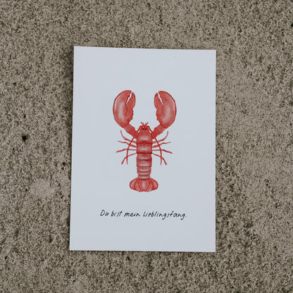 Postkarte Hummer von saltwaterdesigns liegend auf hellem Sanduntergrund.
