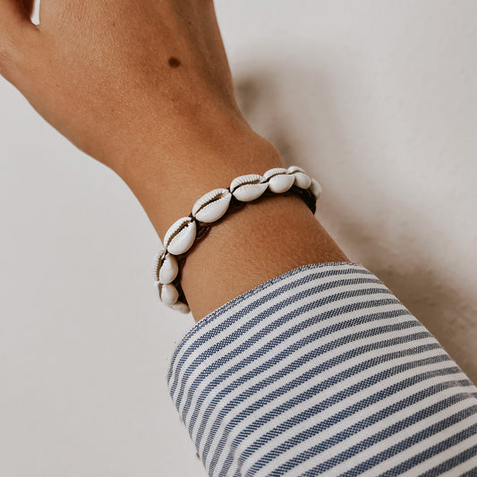 Armband aus weißen Kauri-Muscheln auf dunkelbraunem Band, getragen am Handgelenk vor hellem Hintergrund. Schmuck von saltwaterdesigns.