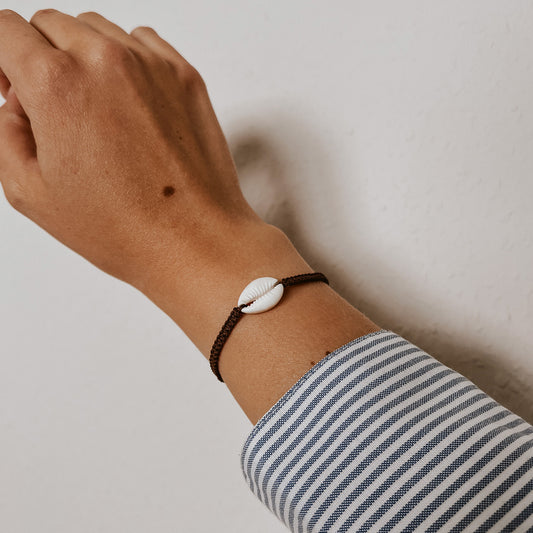Braunes, geflochtenes Armband aus mit einer weißen Kauri Muschel verziert getragen an einem Handgelenk. Armband von saltwaterdesigns.