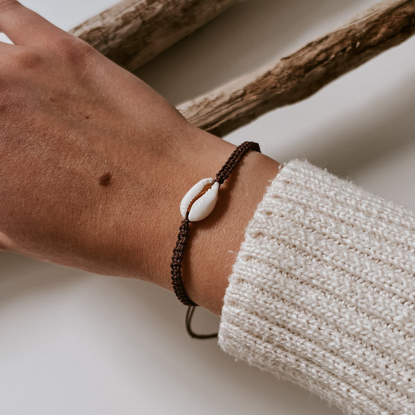 Braunes, geflochtenes Armband aus mit einer weißen Kauri Muschel verziert getragen an einem Handgelenk. Armband von saltwaterdesigns in der Trendfarbe braun.