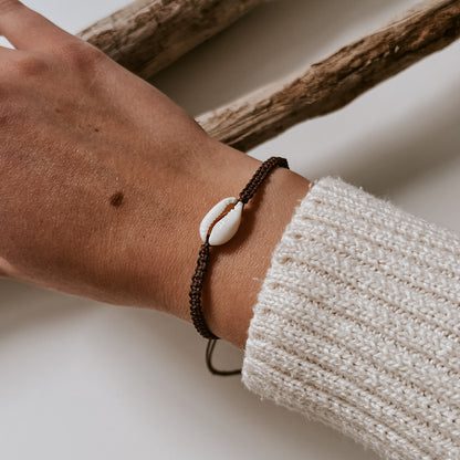 Braunes, geflochtenes Armband aus mit einer weißen Kauri Muschel verziert getragen an einem Handgelenk. Armband von saltwaterdesigns in der Trendfarbe braun.