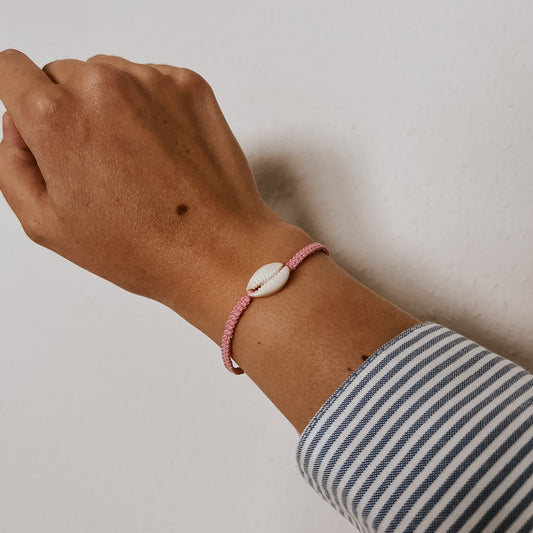 Armband aus weißen Kauri-Muscheln auf zartrosa Band, getragen am Handgelenk vor schlichtem Hintergrund. Boho-Schmuck von Saltwaterdesigns mit sanften Küstenfarben.