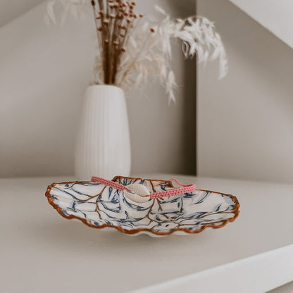 Kauri Muschelarmband in Rosa, präsentiert auf der Schmuckschale Muriwai von saltwaterdesigns. Auf weißem Schrank arrangiert, mit Blumen im Hintergrund – als Deko-Beispiel inszeniert.