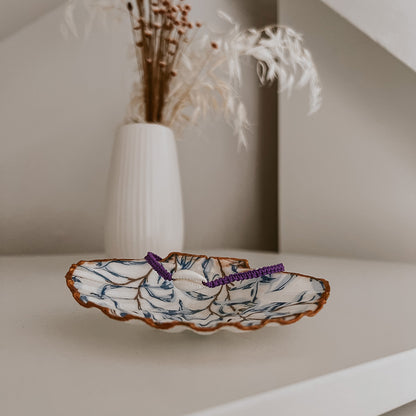 Lila Bali Muschelarmband, dekorativ platziert auf der Schmuckschale Muriwai von saltwaterdesigns. Die Szene auf einem weißen Schrank, umgeben von Blumen, zeigt eine natürliche und stilvolle Deko-Inspiration.