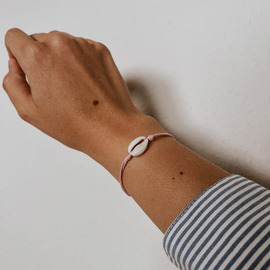Zartes Ocean Armband aus einer weißen Kauri-Muschel auf rosa Band, getragen am Handgelenk vor hellem Hintergrund. Handgefertigter Schmuck von Saltwaterdesigns mit maritimem, eleganten Flair.