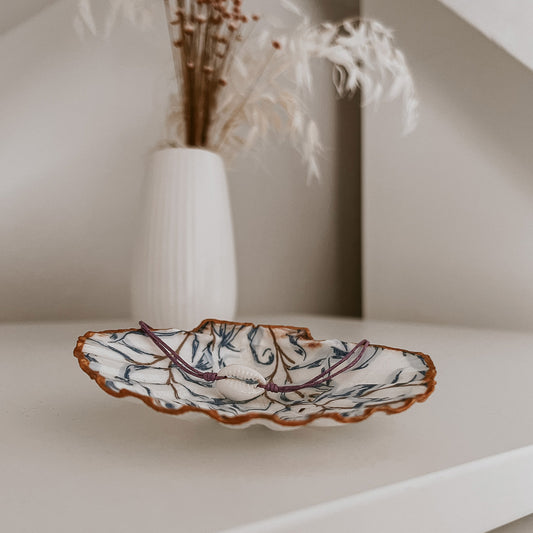 Zartes Ocean Kauri Muschelarmband in Lila, zentral auf der Schmuckschale Muriwai von saltwaterdesigns platziert. Vor einem hellen Schrank und umgeben von Blumen zeigt das Arrangement eine dezente, elegante Deko-Inspiration mit maritimem Flair.