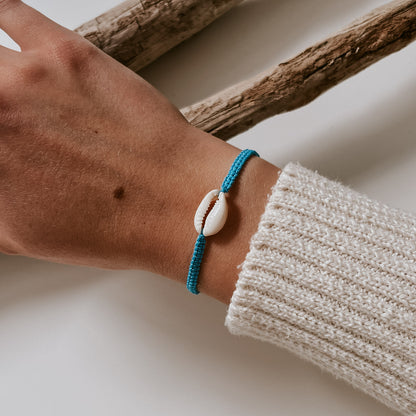 Türkis-farbenes oder hellblaues, geflochtenes Armband mit einer weißen Kauri Muschel in der Mitte verziert, getragen an einem Handgelenk. Armband von saltwaterdesigns.