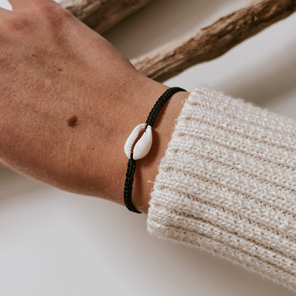 Schwarzes, geflochtenes Armband mit einer weißen Kauri Muschel in der Mitte verziert, getragen an einem Handgelenk. Armband von saltwaterdesigns.
