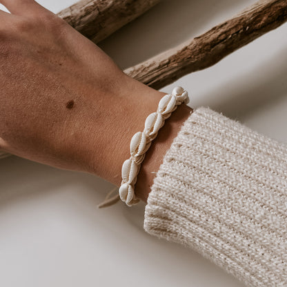 Armband aus weißen Kauri-Muscheln auf weißem Band, getragen am Handgelenk vor hellem Hintergrund mit Strandholz Ambiente. Schmuck von saltwaterdesigns.