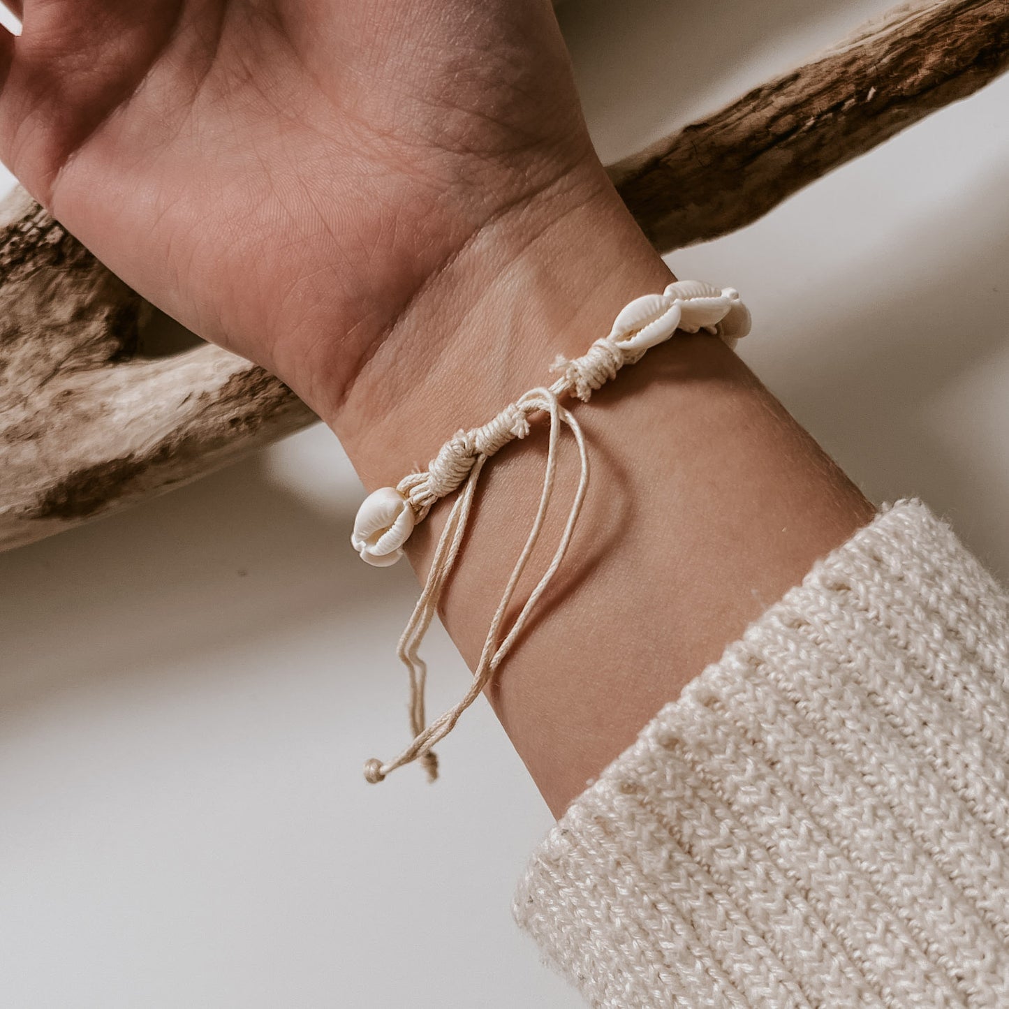 Ansicht von oben mit dem Armband weißen Kauri-Muscheln auf weißem Band, getragen am Handgelenk vor hellem Hintergrund mit Strandholz Ambiente. Schmuck von saltwaterdesigns.