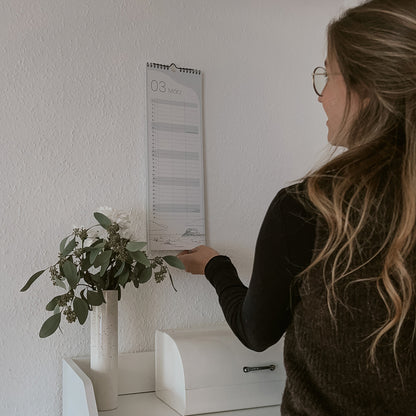 Frau steht vorm Küchenregal und klappt Klanederblatt um vom schmalen Kalender 2026 von saltwaterdesigns mit Illustration von Sankt Peter Ording.