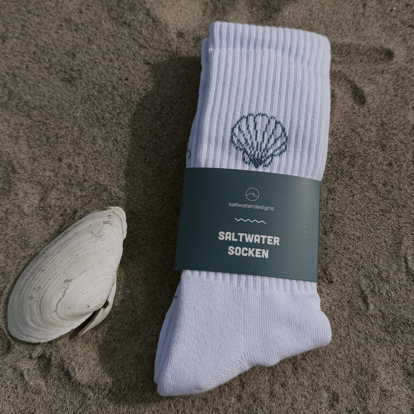 Saltwater Socken Muschel von saltwaterdesigns, weiß gefaltete Tennissocke mit Muscheldesign und petrolblauer Banderole auf Sand liegend.