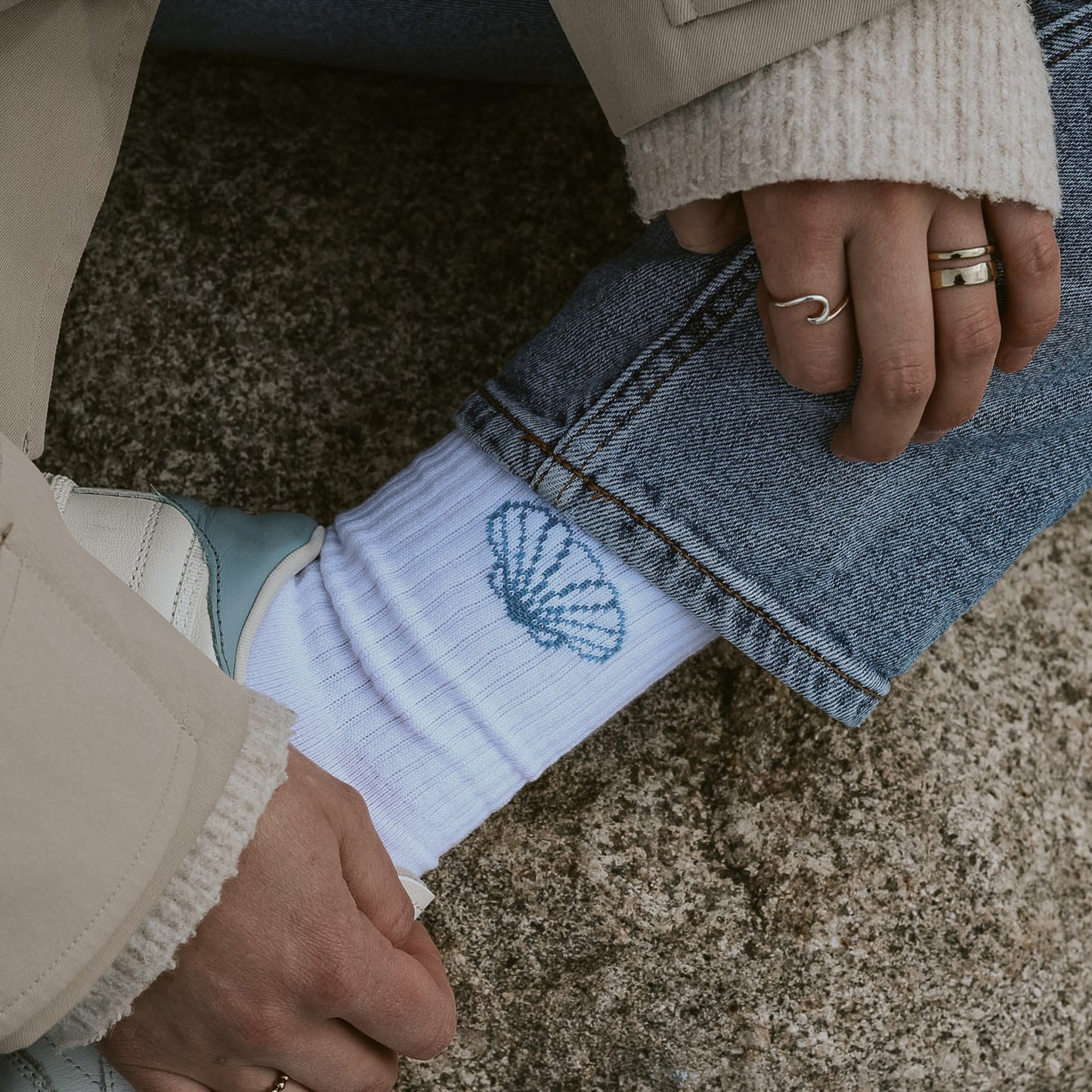 Nahaufnahme Saltwater Socken mit eingestrickter Jakobsmuschel, kombiniert mit Jeans und weißen Sneakern am Strand.